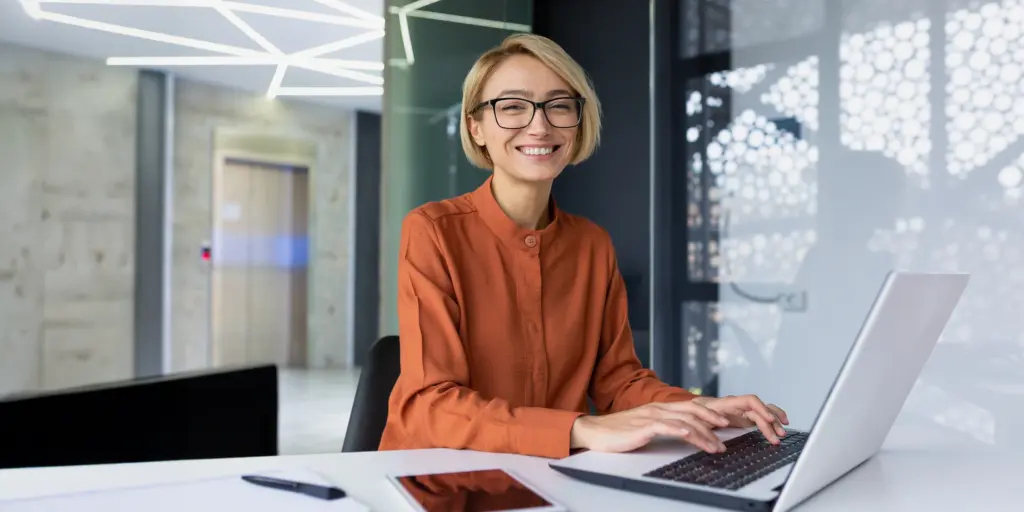 Frau in modernem Büro, lächelt am Laptop, trägt orangefarbene Bluse, Tablet daneben, helle Beleuchtung.