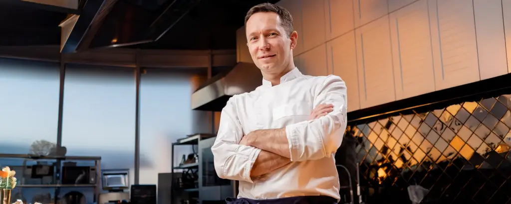 Ein lächelnder Koch in weißer Jacke steht in einer modernen, professionellen Küche mit verschränkten Armen.
