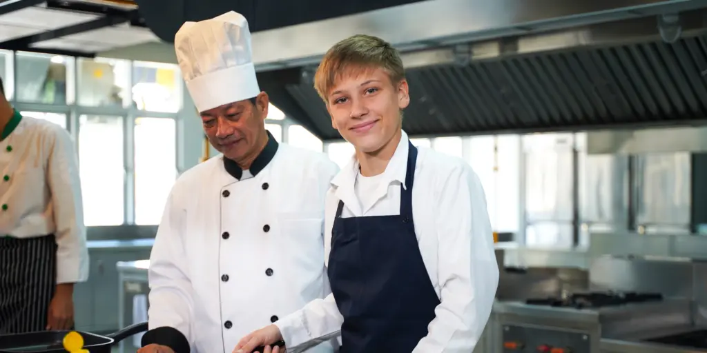 Zwei Köche, ein Erwachsener und ein Junge, lächeln in einer modernen Restaurantküche.