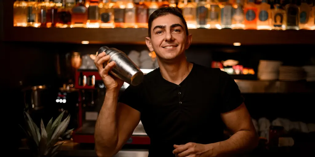 Ein Barkeeper lächelt und schüttelt einen Cocktailmixer vor einer Bar mit vielen Flaschen im Hintergrund.